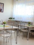 Wnętrze Domu Pomocy Społecznej. Trzy kremowe stoły na stalowych nogach. Na blatach stołów kompozycje z kwiatów. Na kremowych ścianach dwa obrazy. W tle okno przesłonięte ażurową firaną.